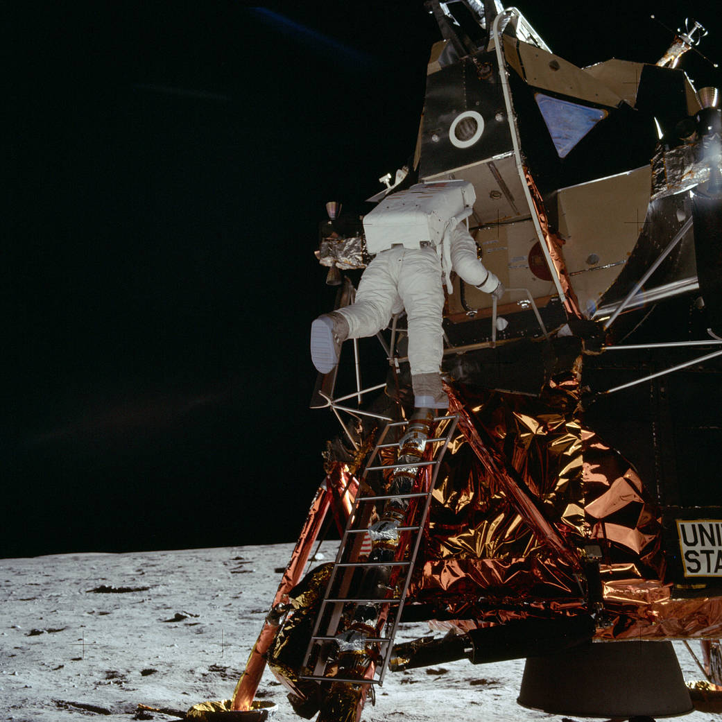 Buzz Aldrin climbing down the ladder of the lunar module Eagle. The lunar surface is brightly lit; the sky behind the module is pure black, with no stars visible.
