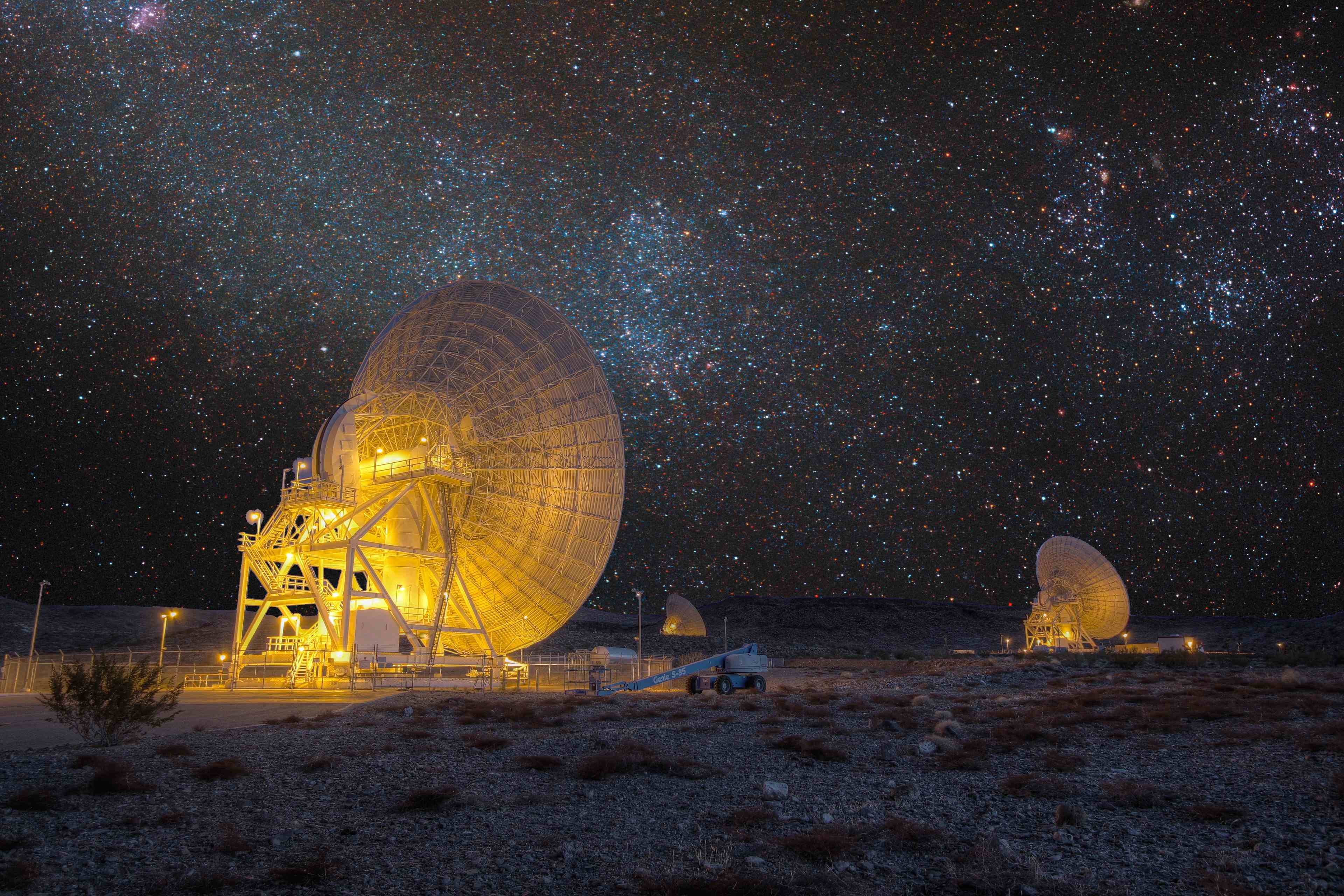A large gold-lit radio dish at the Goldstone Deep Space Communications Complex's Apollo Valley at night, a second dish visible in the distance, under a dense field of stars with visible clusters and nebulae.