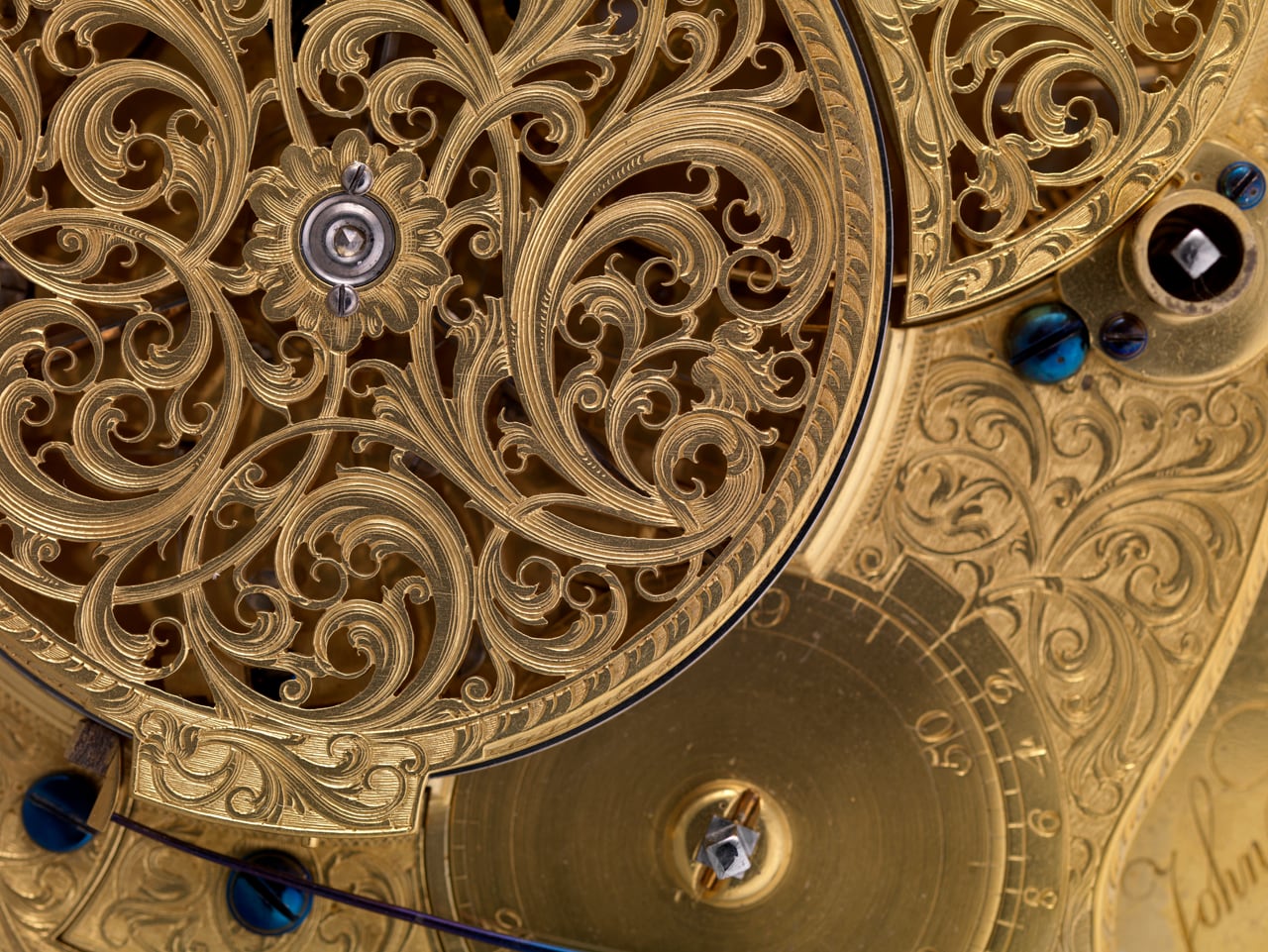 A close view of the inside of the watch: gilded brass plates pierced and engraved edge to edge with dense scrolling foliage, a rosette at the centre, the steelwork blued, every surface a wheel does not touch carved by hand.